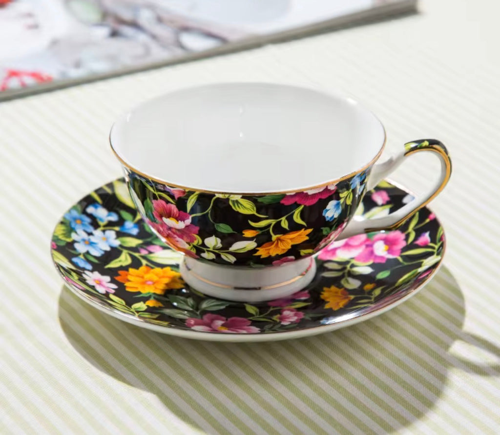 A floral teacup and saucer set on a striped tablecloth. The cup and saucer feature a black background with a vibrant multicolor floral pattern in pink, orange, yellow, blue, and green, accented with gold trim and a white interior.