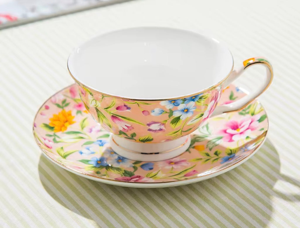 A floral teacup and saucer set on a striped tablecloth. The cup and saucer feature a light peach-pink base decorated with a colorful floral pattern of pink, yellow, blue, and purple flowers with green leaves, accented by gold trim and a white interior.