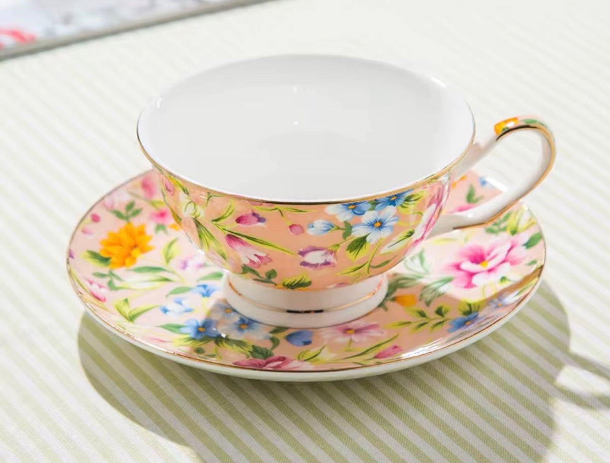 A floral teacup and saucer set on a striped tablecloth. The cup and saucer feature a light peach-pink base decorated with a colorful floral pattern of pink, yellow, blue, and purple flowers with green leaves, accented by gold trim and a white interior.