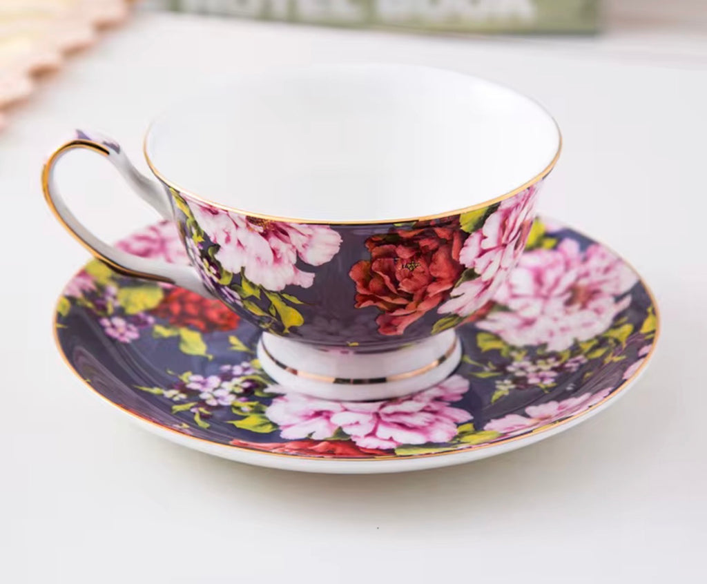 A floral porcelain teacup and saucer set on a light surface. The cup and saucer feature a dark navy blue background with large pink and red flowers, green leaves, and gold trim, including a gold rim and handle.