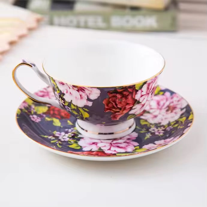 A floral porcelain teacup and saucer set on a white surface, featuring a dark purple background with pink, red, and white flowers and gold trim.