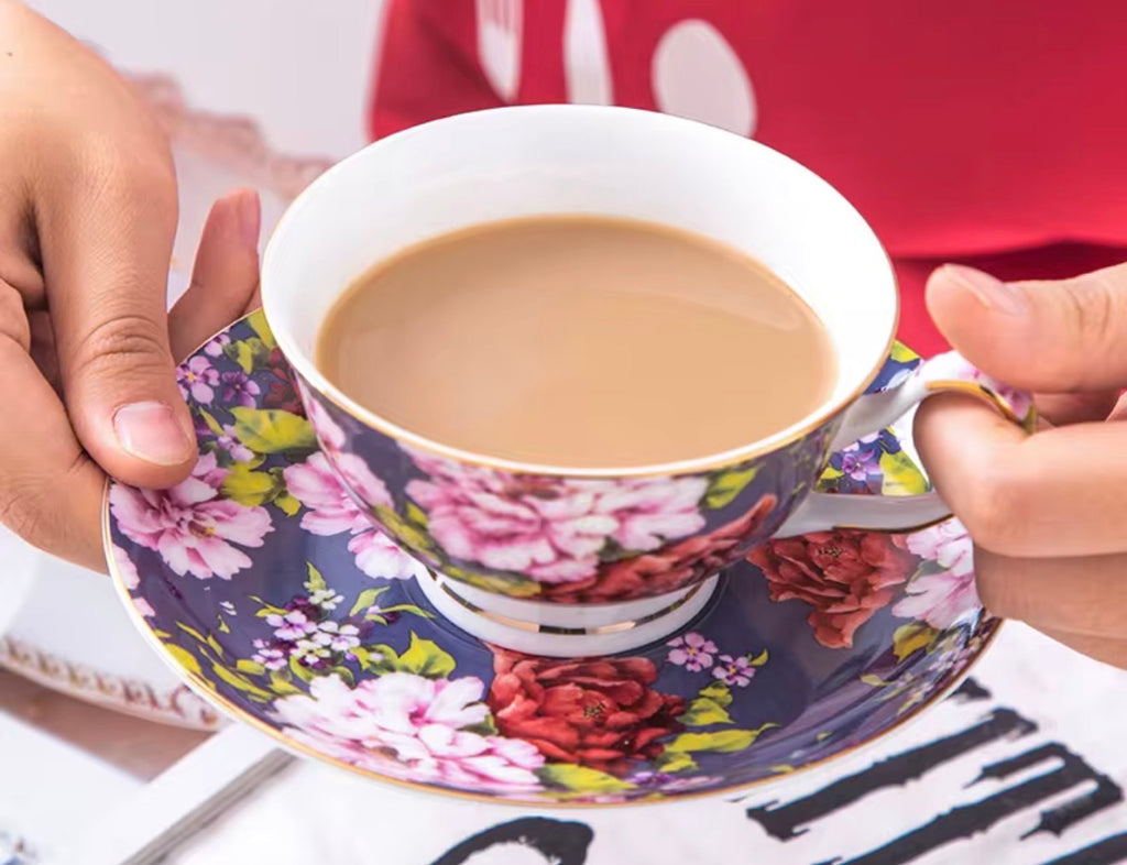 A close-up of a person holding a floral teacup and saucer filled with milk tea. The cup and saucer feature a dark navy blue background with large pink and red flowers, green leaves, and gold trim.