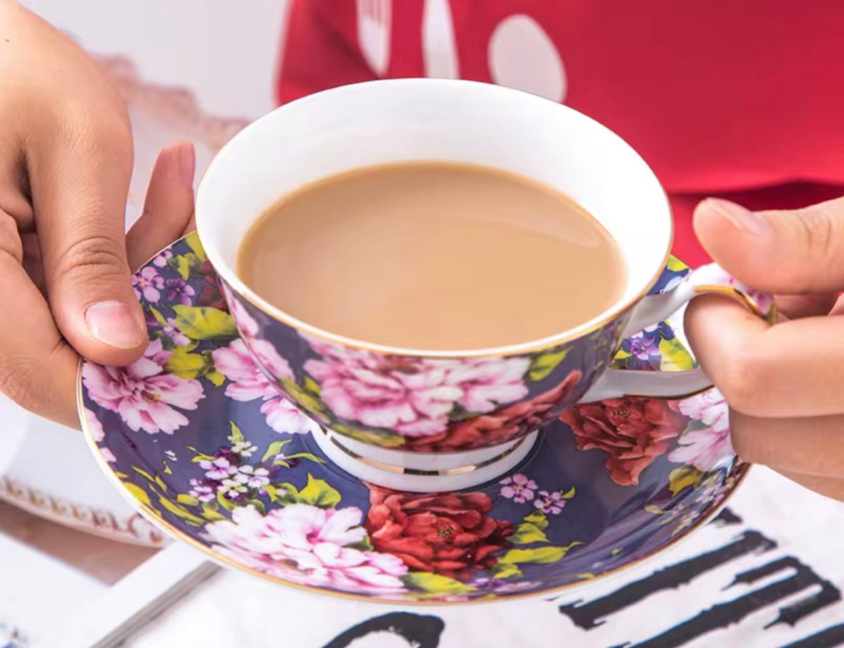 A close-up of a person holding a floral teacup and saucer filled with milk tea. The cup and saucer feature a dark navy blue background with large pink and red flowers, green leaves, and gold trim.