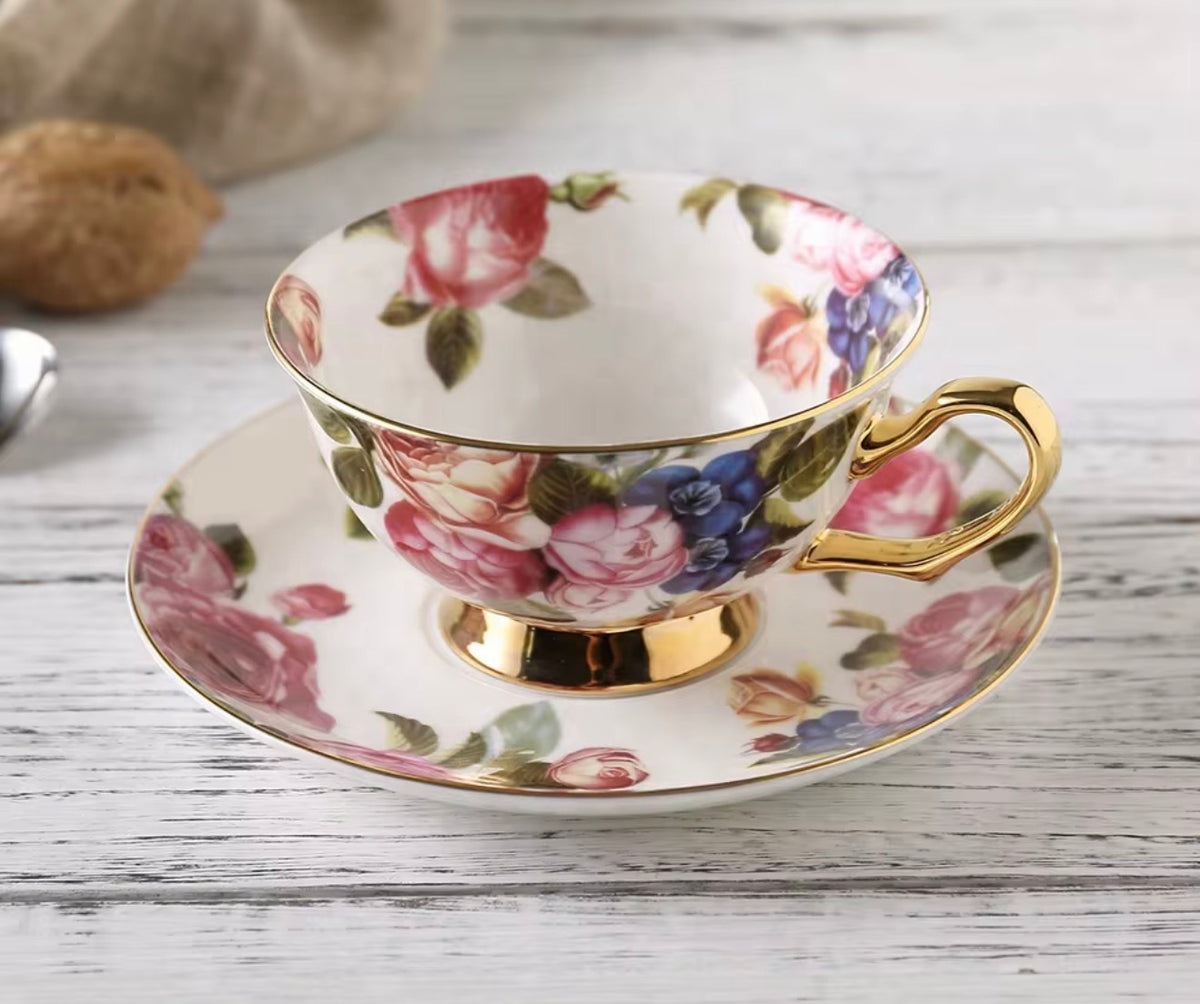 A floral porcelain teacup and saucer set with gold trim, featuring pink and blue flowers on a white background, styled on a rustic white wooden surface.