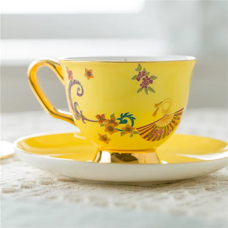 A yellow ceramic teacup with a matching saucer, featuring hand-painted floral and bird motifs with gold trim, set on a light textured surface.