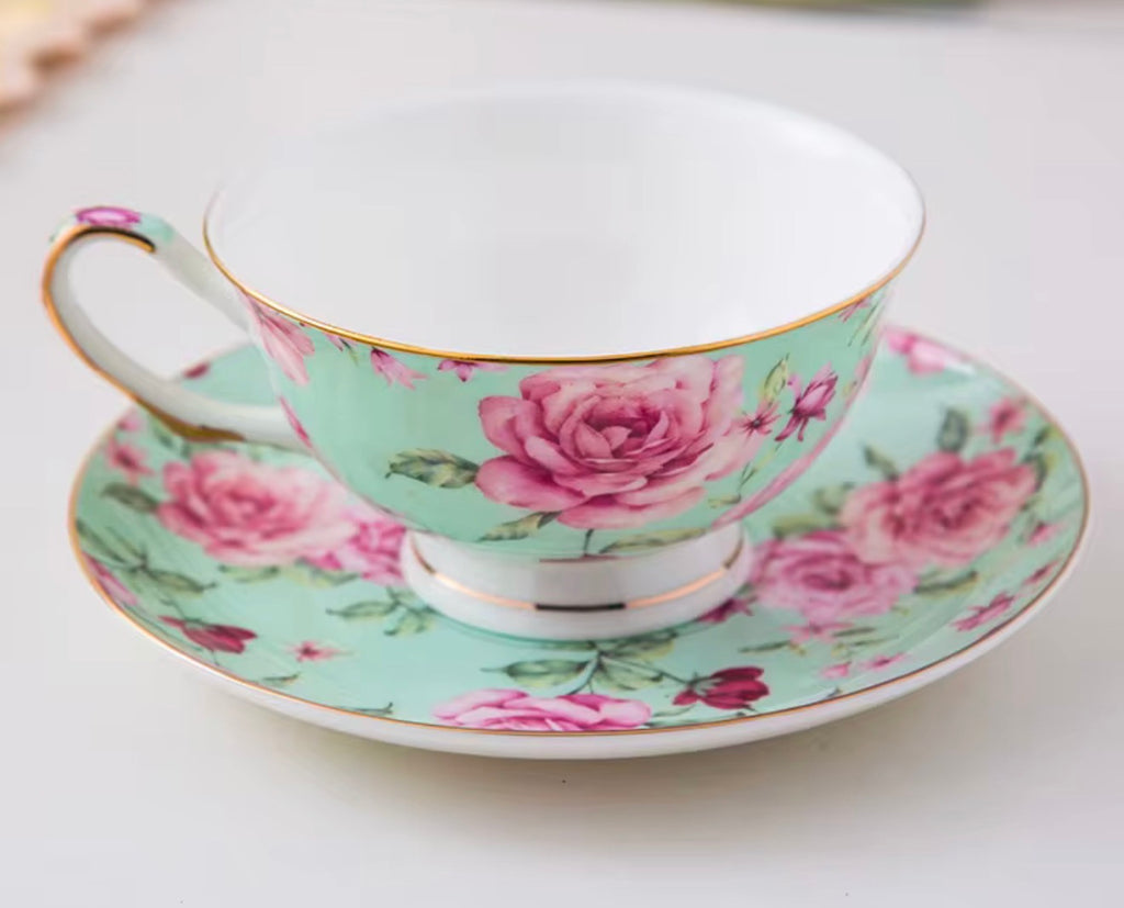 A porcelain teacup and saucer set decorated with a pink rose floral pattern on a mint green background, accented with gold trim and a white interior.